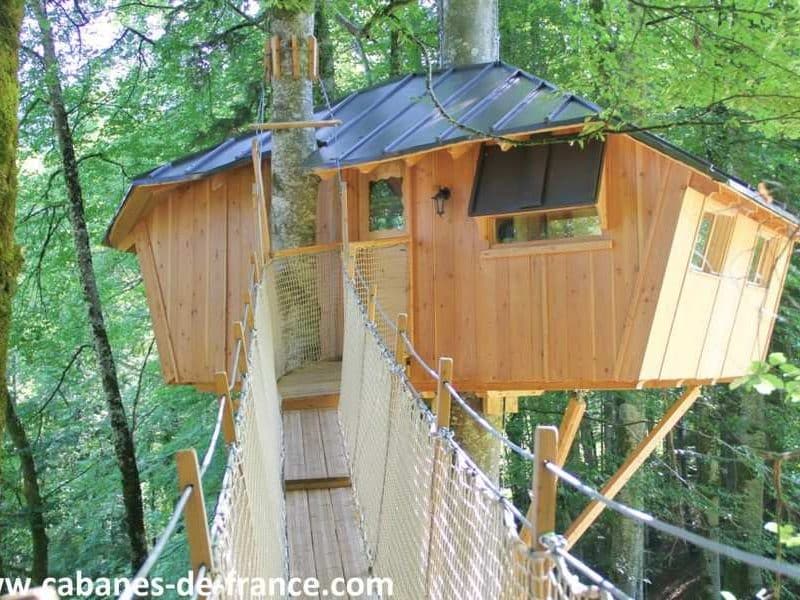 Cabane perchée en bois dans les arbres, accessible par un pont suspendu.