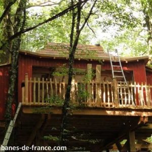 Cabane perchée en bois rouge, entourée darbres, avec une terrasse accueillante.
