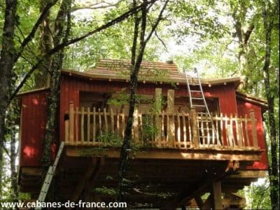 Cabane perchée en bois rouge, entourée darbres, avec une terrasse accueillante.