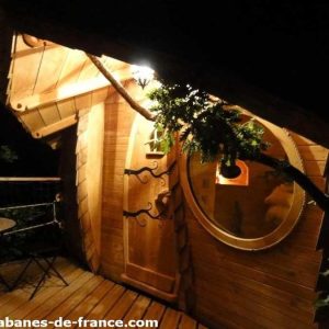 Cabane en bois originale, éclairée la nuit, avec une porte ronde et terrasse en bois.