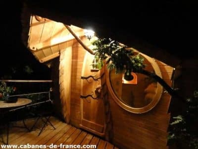 Cabane en bois originale, éclairée la nuit, avec une porte ronde et terrasse en bois.
