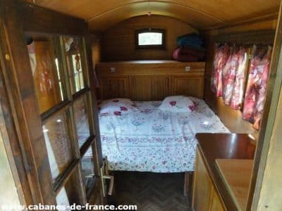 Intérieur dune roulotte en bois, lit douillet avec draps fleuris et fenêtres lumineuses.