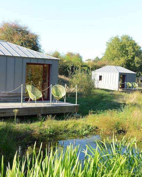 Cabane moderne en bois avec chaises vertes, entourée de verdure et deau.