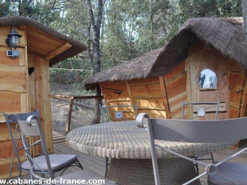 Cabane en bois avec toit de chaume, terrasse en bois et mobilier extérieur.