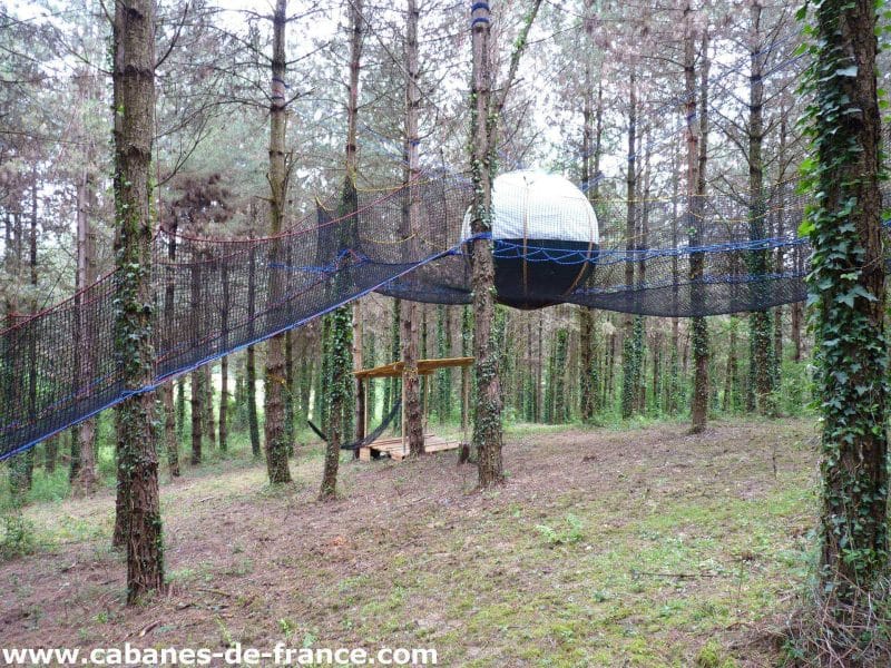 Cabane suspendue dans les arbres, avec un espace de détente en filet entre les troncs.