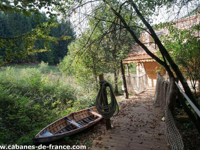 Cabane perchée dans les arbres, accès par un chemin en bois entouré de verdure.