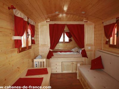 Hébergement insolite : roulotte en bois avec rideaux rouges et lit douillet.