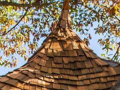 Cabane perchée en bois avec un toit en ardoise, entourée darbres verdoyants.