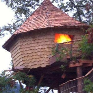 Cabane perchée en bois dans les arbres, avec un toit en ardoise et une lumière chaleureuse.