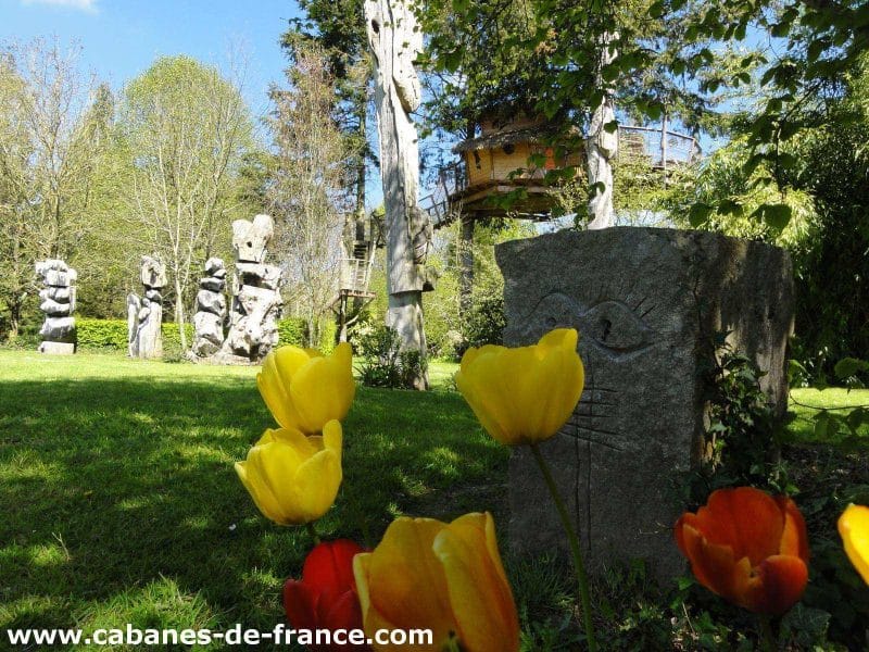 Cabane dans les arbres entourée de sculptures et de fleurs colorées.