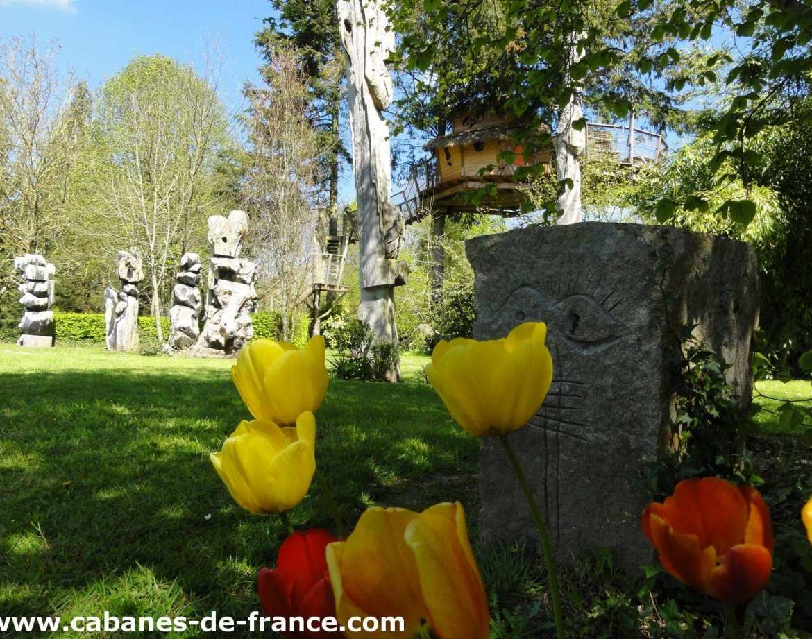 Cabane perchée en Bretagne, entourée de sculptures et de fleurs colorées.