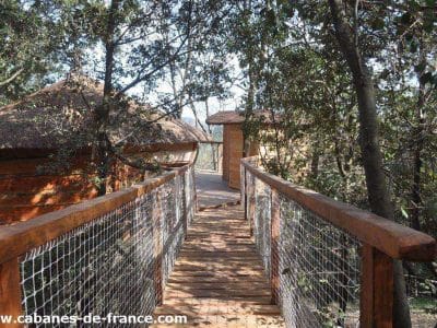 Cabane perchée en bois, accessible par un pont suspendu, entourée de verdure.