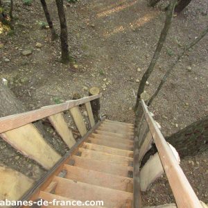 Cabane perchée en bois, escaliers menant à un cadre naturel verdoyant.
