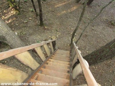 Cabane perchée en bois, escaliers menant à un cadre naturel verdoyant.