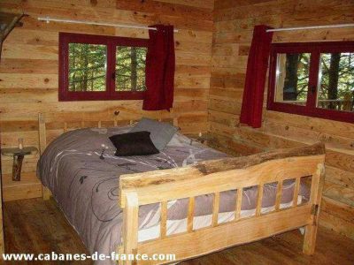Cabane en bois dans les arbres, avec lit en bois et fenêtres rouges.