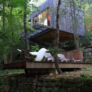 Cabane moderne suspendue, entourée darbres, avec terrasse en bois et mobilier design.