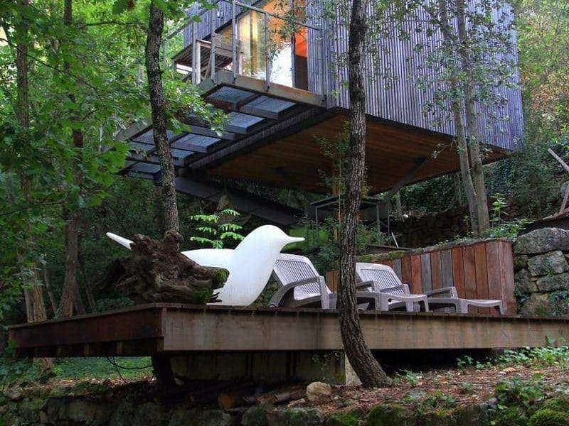 Cabane suspendue en bois, entourée de verdure, avec terrasse en bois et mobilier moderne.