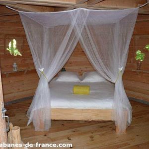 Cabane en bois avec lit double, drapée de voilages légers et fenêtres colorées.