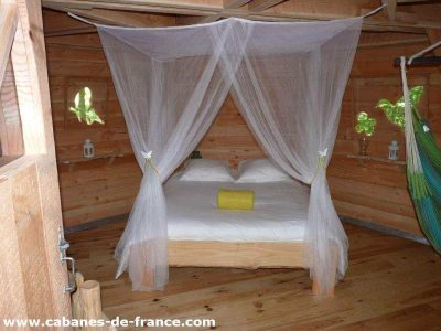Cabane en bois avec lit double, drapée de voilages légers et fenêtres colorées.