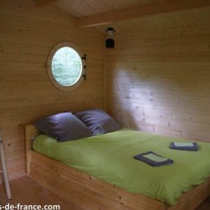 Cabane en bois à Midi-Pyrénées, lit douillet avec draps verts et fenêtre ronde.