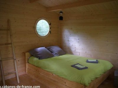 Cabane en bois à Midi-Pyrénées, lit douillet avec draps verts et fenêtre ronde.