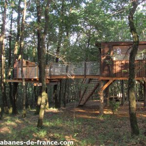 Cabane perchée dans les arbres, entourée de verdure, avec terrasse en bois.