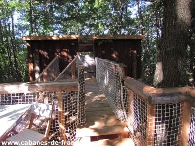 Cabane perchée dans les arbres, accessible par un pont en bois et filet.