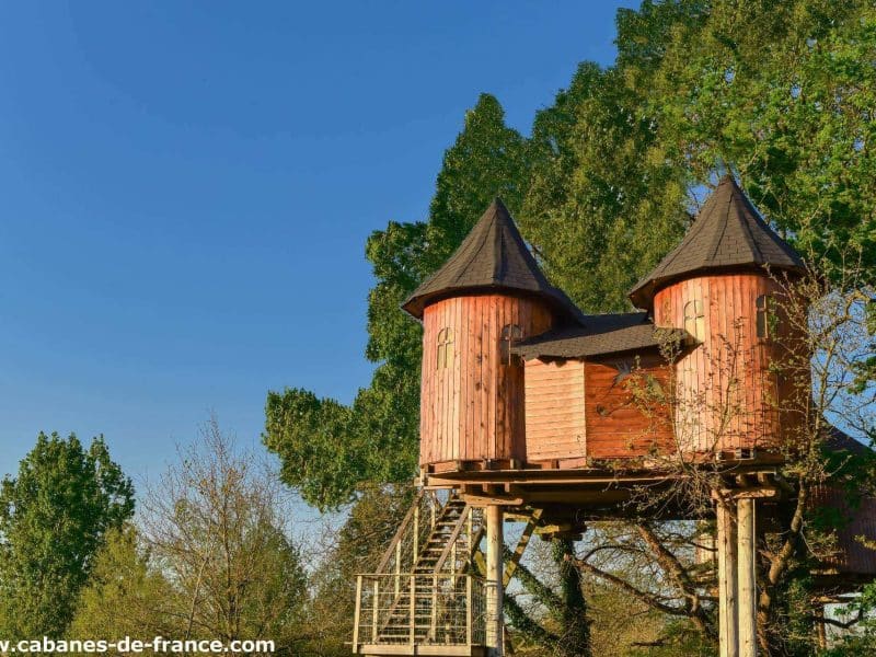 Cabane perchée en bois avec deux tourelles, entourée darbres verdoyants.