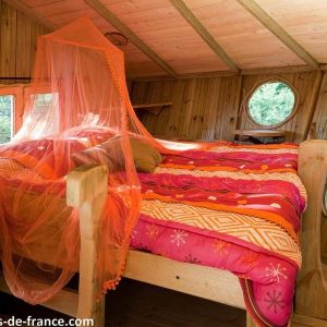Cabane perchée en bois avec un lit coloré et un voile en tulle, ambiance chaleureuse.