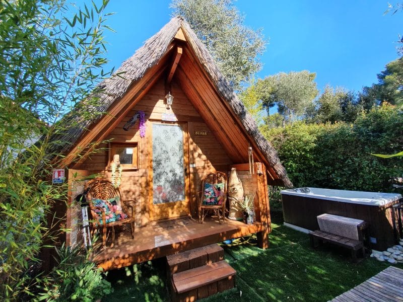 Cabane en bois avec toit de chaume, jacuzzi et jardin verdoyant à proximité.