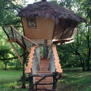 Cabane perchée en bois avec toit de chaume, accessible par un pont en bois.