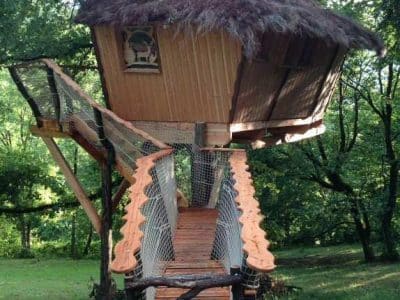 Cabane perchée en bois avec toit de chaume, accessible par un pont en bois.