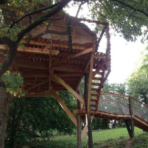 Cabane perchée en bois, accessible par un pont, entourée darbres verdoyants.
