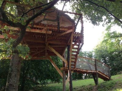 Cabane perchée en bois, accessible par un pont, entourée darbres verdoyants.