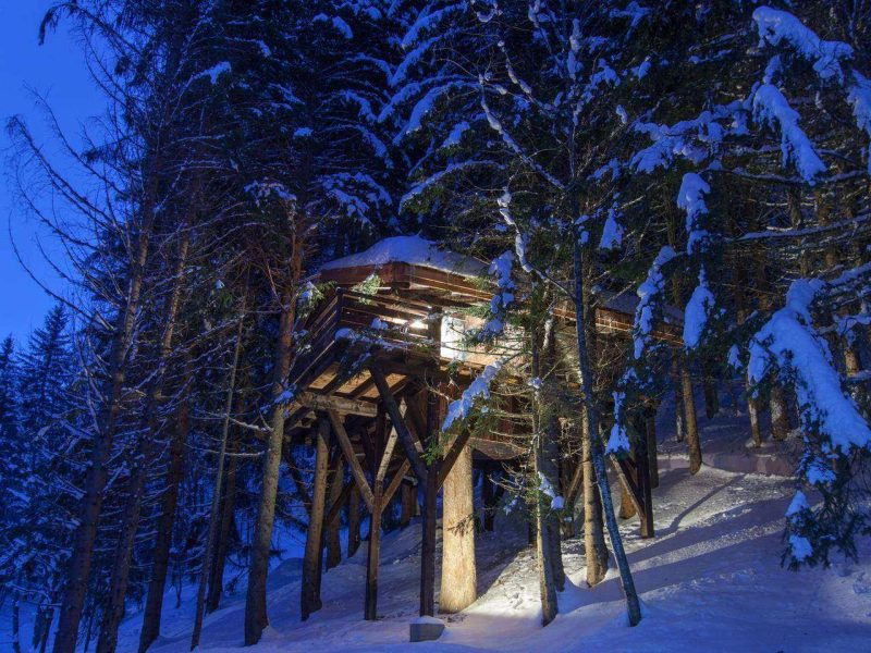 Cabane perchée en bois, illuminée, entourée de sapins sous la neige en Auvergne-Rhône-Alpes.