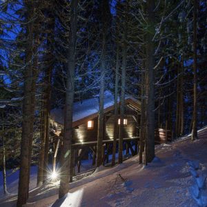 Cabane perchée en bois, illuminée, entourée de sapins sous la neige en Auvergne-Rhône-Alpes.