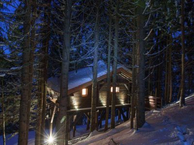 Cabane perchée en bois, illuminée, entourée de sapins sous la neige en Auvergne-Rhône-Alpes.