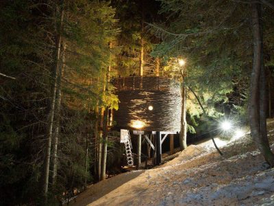 Cabane perchée dans les arbres, illuminée, entourée de neige et de sapins en Auvergne.