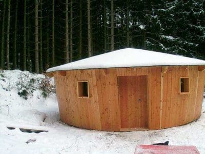 Hébergement insolite en bois, cabane ronde sous la neige, entourée de sapins.