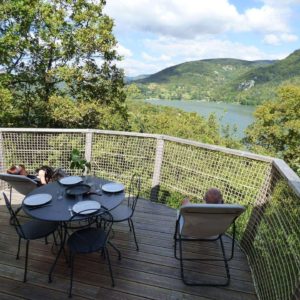 Chalet en bois avec terrasse panoramique sur un lac et des montagnes verdoyantes.