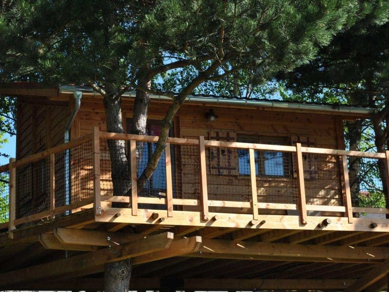 Cabane perchée en bois, entourée darbres, avec une terrasse en bois spacieuse.