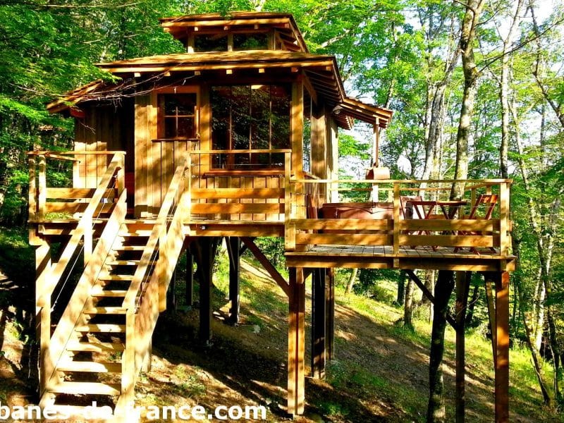 Cabane dans les arbres en bois, perchée sur pilotis, entourée de verdure.