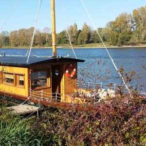 Hébergement insolite : une péniche en bois amarrée au bord de la rivière, entourée de verdure.