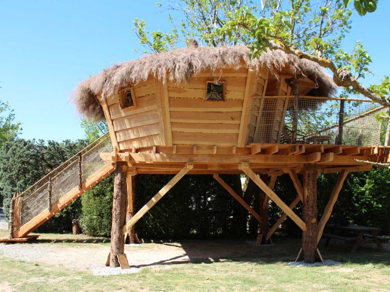Cabane perchée en bois avec toit en chaume, entourée de verdure.