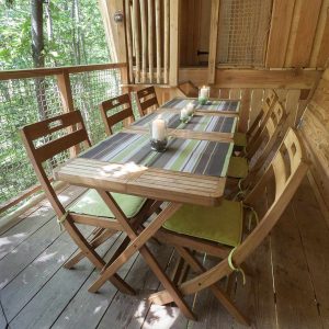 Cabane perchée en bois avec une table en bois et des chaises, entourée de verdure.
