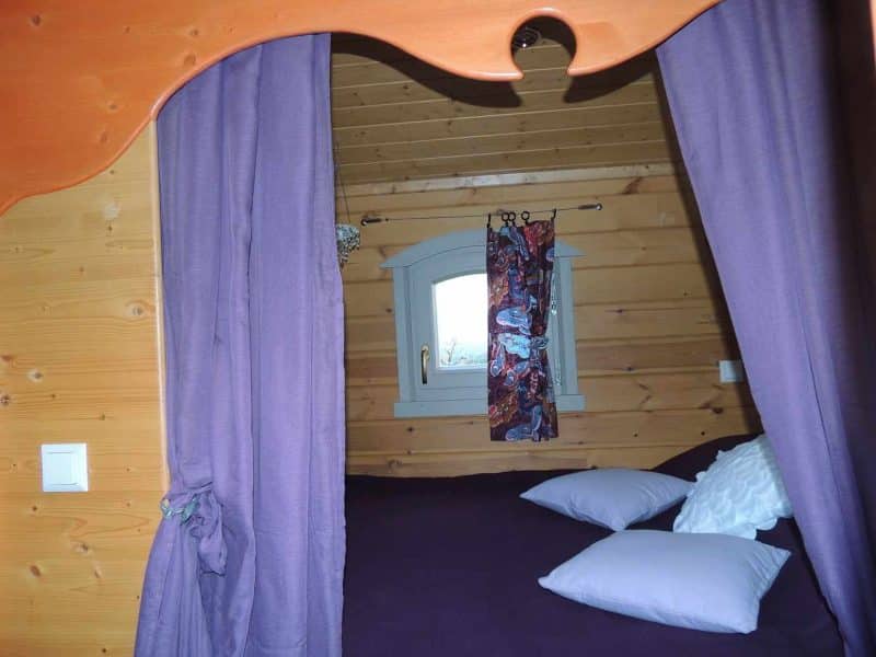 Chalet cosy avec lit douillet, rideaux violets et fenêtre lumineuse.