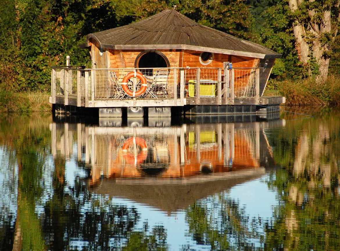 Cabane flottante en bois sur un lac, entourée de verdure et reflet paisible.