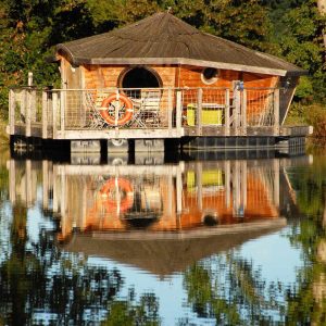 Cabane flottante en bois sur un lac, entourée de verdure et reflet paisible.