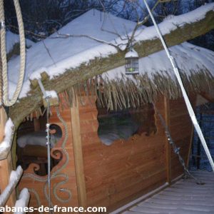 Cabane perchée en bois sous la neige, avec un toit en chaume et une vue sur la nature.