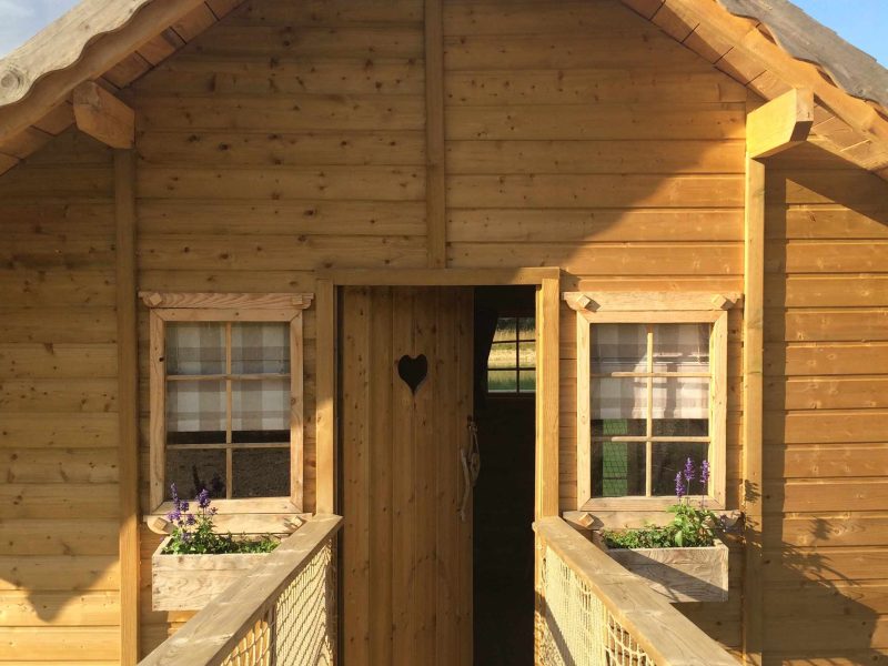 Cabane en bois avec un cœur découpé à la porte, entourée de fleurs.